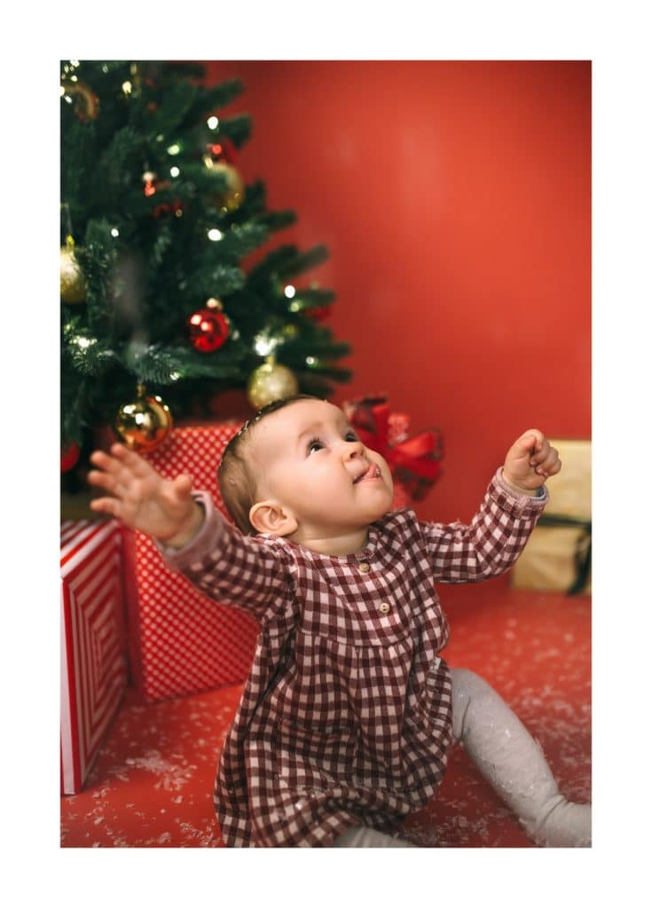 Babybilder zu Weihnachten. Mädchen im roten Kleid sitzt vor dem geschmückten Weihnachtsbaum.